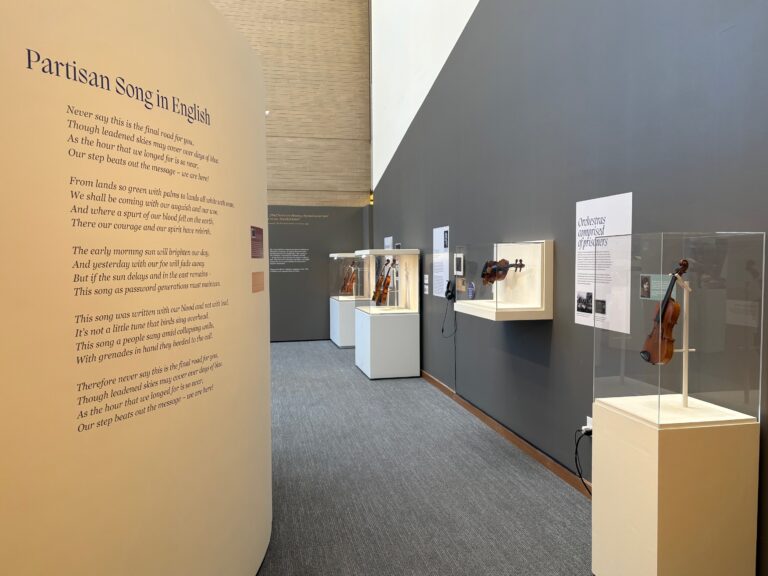 Museum exhibition gallery featuring violins displayed in individual glass cases on white pedestals. A large wall panel on the left displays the text "Partisan Song in English" with several stanzas of lyrics. The violins are arranged along gray walls with accompanying informational plaques.