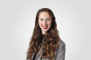 Sam Goldberg, HERC's Education Director, smiles at the camera against a light gray background. She has long, wavy brown hair and wears red lipstick with a gray patterned sweater.