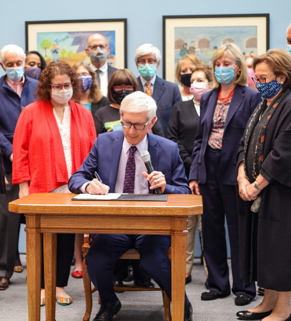 Wisconsin Governor Tony Evers, dressed in a navy suit and purple tie, sits at a wooden desk signing Act 30 into law while holding a microphone. A diverse group of approximately fifteen masked supporters stands behind him in a room with light blue walls and framed artwork. Representatives from the Nathan and Esther Pelz Holocaust Education Resource Center were among those present, having played a key role in advocating for the bill's passage