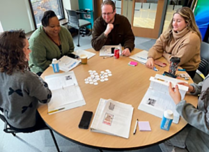 A diverse group of adults participates in a Holocaust education workshop, seated around a round table with open workbooks, printed materials, and discussion resources spread across the surface.