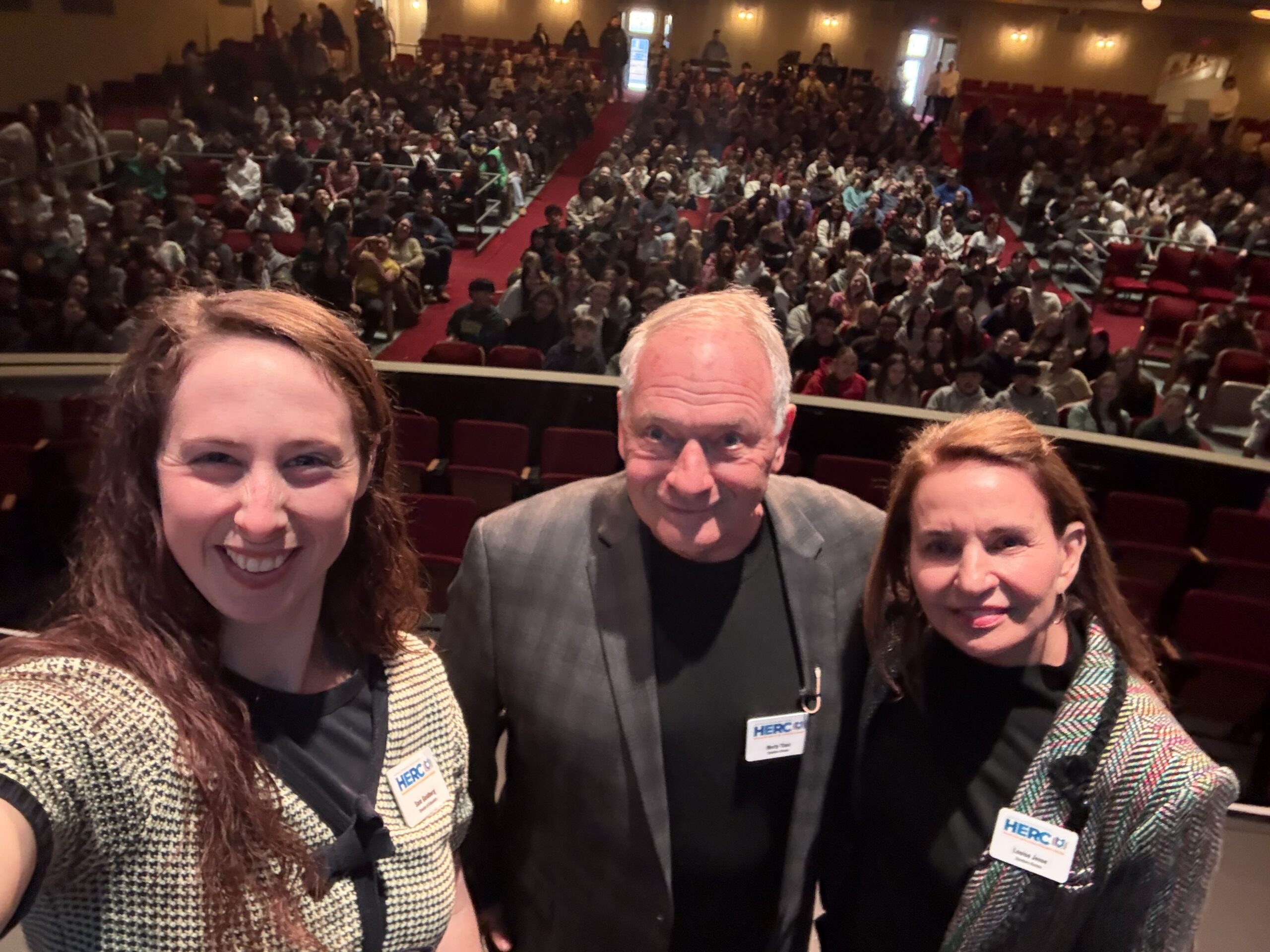Sam Goldberg, Marty Thau, and Louise Jesse smile before a packed auditorium of students at a HERC program.