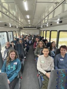 Students and chaperones smile for a photo aboard a charter bus en route to the Illinois Holocaust Museum & Education Center. Several participants wear name tags, and green grass is visible through the bus windows.