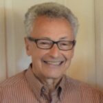 An older man, Holocaust survivor Werner Richheimer, with short gray hair and dark rectangular-framed glasses smiles warmly at the camera. He is wearing a striped button-down shirt in muted tones. The background appears to be a light-colored wall or door.