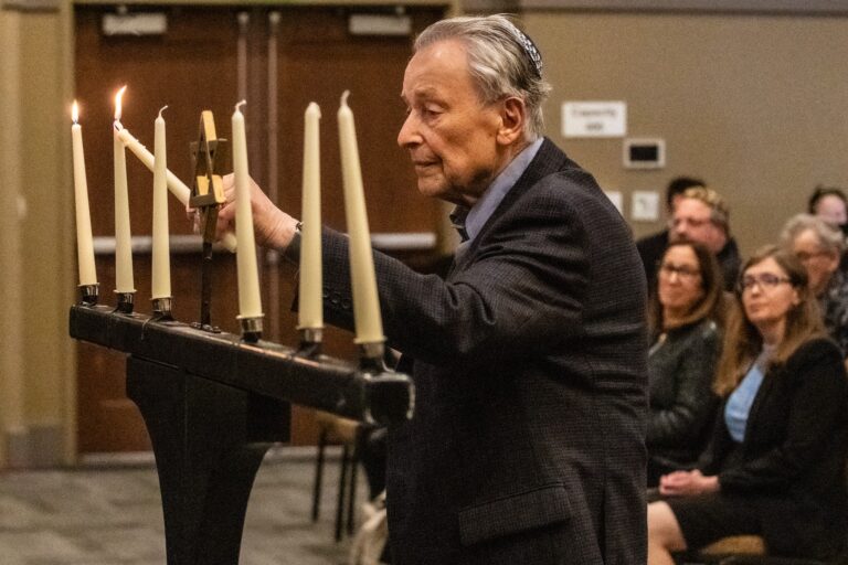 An elderly man, Holocaust survivor Werner Richheimer wearing a kippah and dark blazer lights candles on a large menorah, holding a lit taper to the unlit candles. Several candles are already burning. An audience of adults is seated in the background of the indoor event space.