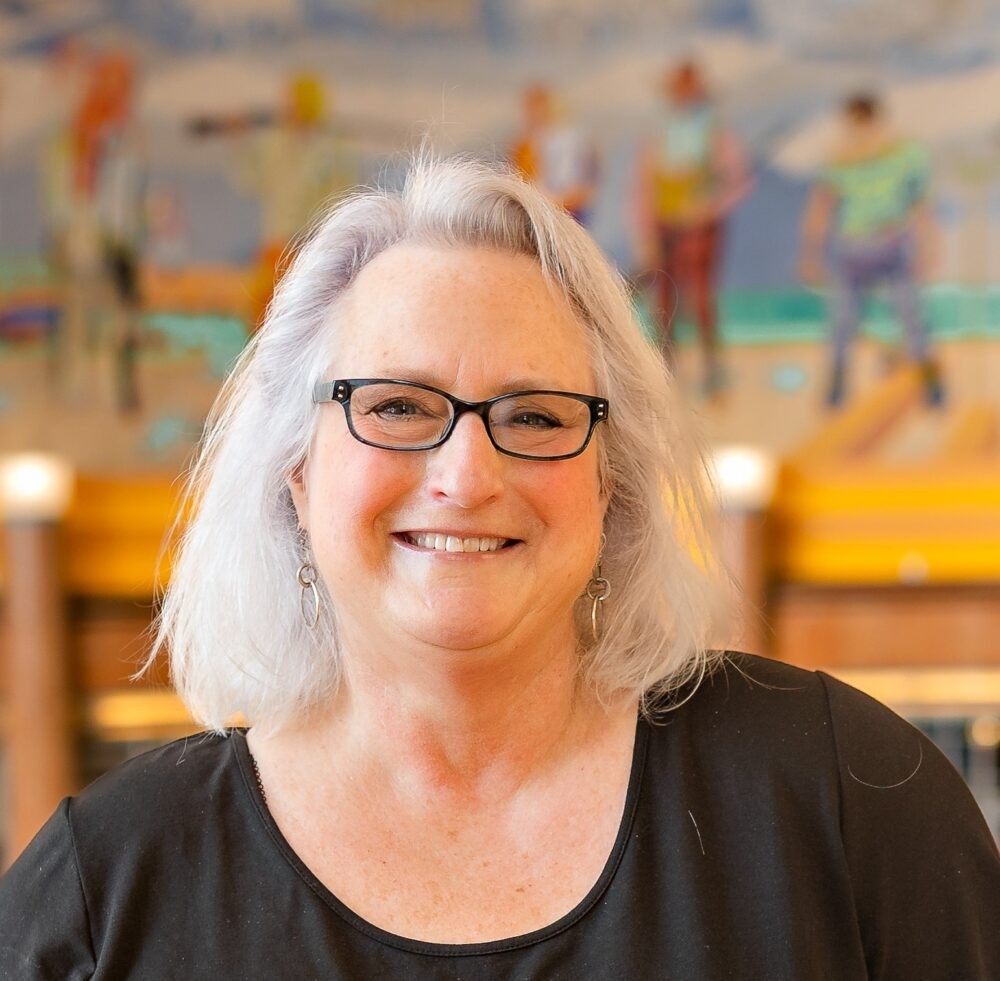 Jodi Elowitz, a speaker for HERC's Navigating the Holocaust Summer Workshop, smiles warmly at the camera. She has shoulder-length silver hair, black rectangular glasses, and wears a black top.