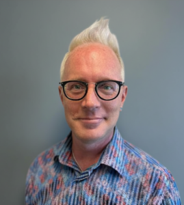 Justin Welch, HERC staff member, smiling headshot against a gray background, wearing glasses and a patterned blue shirt.