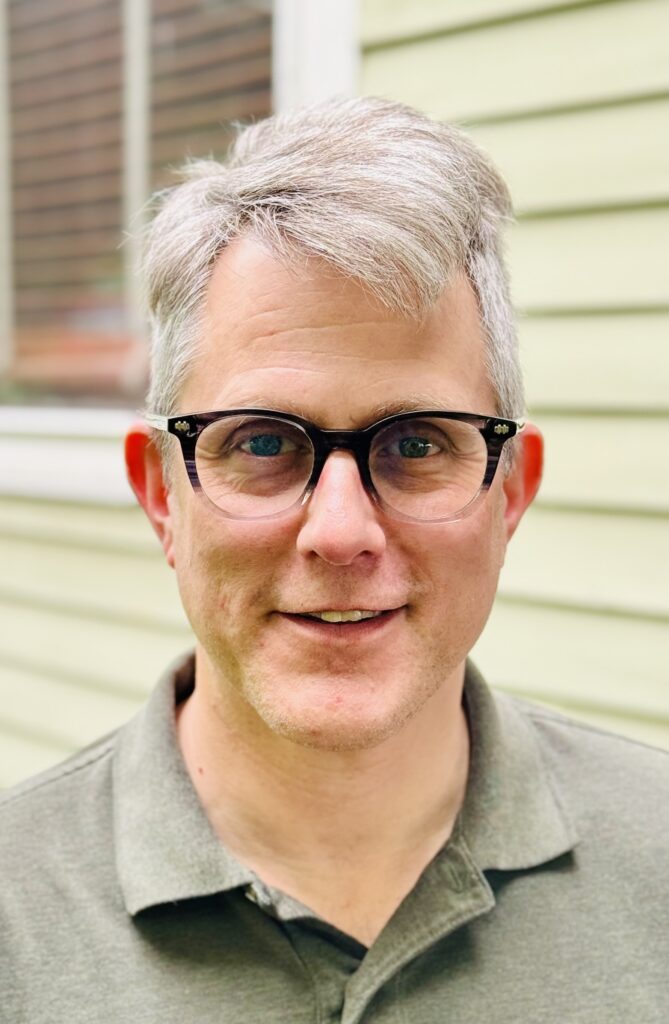 Daniel H. Magilow, a speaker for HERC's Navigating the Holocaust Summer Workshop, smiles at the camera. He has short silver hair, dark tortoiseshell glasses, and wears a gray polo shirt, photographed outdoors.