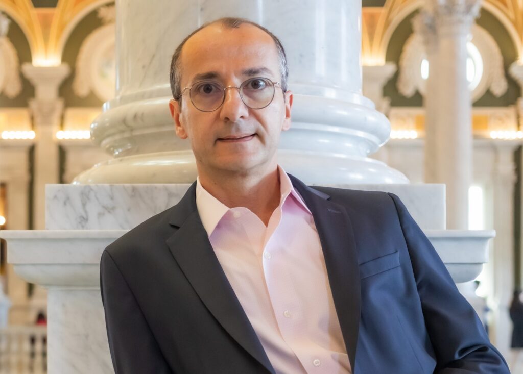 Khatchig Mouradian, a speaker for HERC's Navigating the Holocaust Summer Workshop, leans against a marble surface with hands clasped. He wears round wire-rimmed glasses and a navy blazer over a pink shirt, posed in front of ornate marble columns.