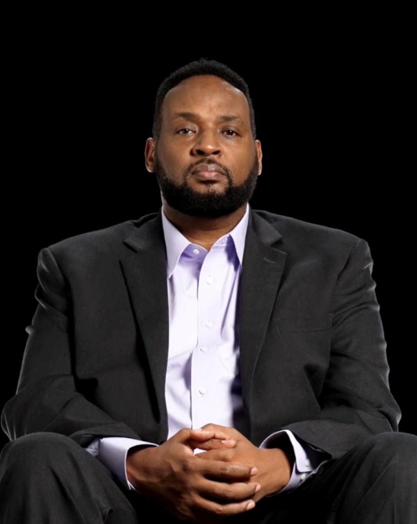 Kizito Kalima, a speaker for HERC's Navigating the Holocaust Summer Workshop, sits with hands clasped and gazes directly at the camera. He wears a dark blazer over a light lavender shirt, photographed against a black background.