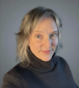 Sara Sillars, Marketing Manager at the Nathan and Esther Pelz Holocaust Education Resource Center, smiles warmly at the camera. She has shoulder-length blonde and gray hair and light eyes, and wears a dark ribbed turtleneck sweater. The photo is taken against a soft gray background.
