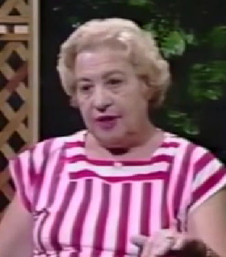 Hannah Noach, a Holocaust survivor, speaks animatedly during a recorded interview. She has short white hair and wears a pink and white striped top.