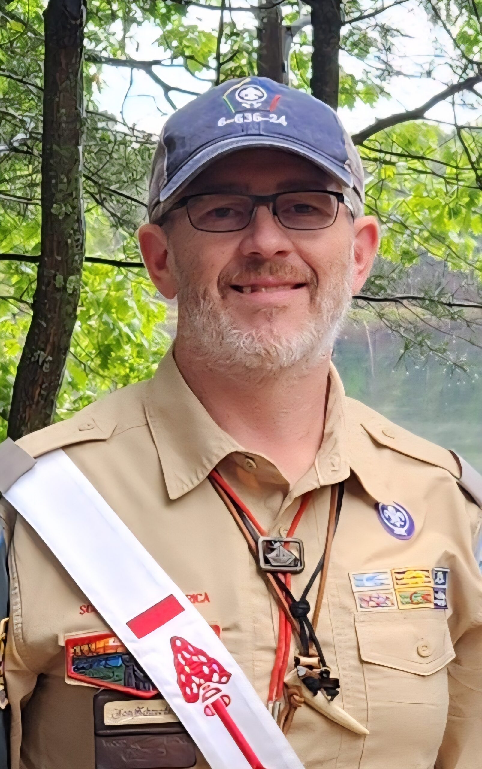 Man with gray beard, baseball cap, and glasses dressed in a tan uniform shirt with badges.